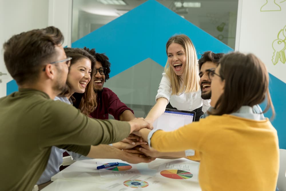 A group of people gather around a table while their hands stacked together showing a positive team culture.